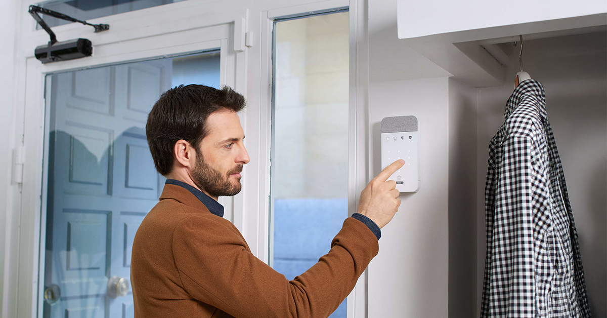 Homem de barba com casaco marrom a ajustar um painel de controlo na parede, utilizando um detetor magnético branco com tecnologia Shock Sensor.
