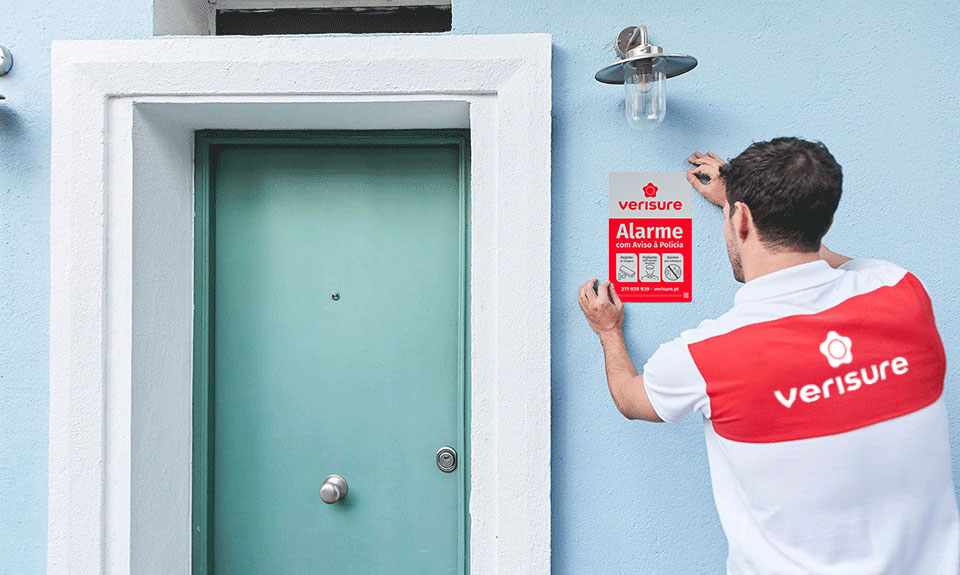 Técnico da Verisure a colocar uma placa de alarme ao lado de uma porta de entrada de uma vivenda, garantindo proteção contra roubos e invasões Técnico da Verisure a colocar uma placa de alarme ao lado de uma porta de entrada de uma vivenda, garantindo proteção contra roubos e invasões